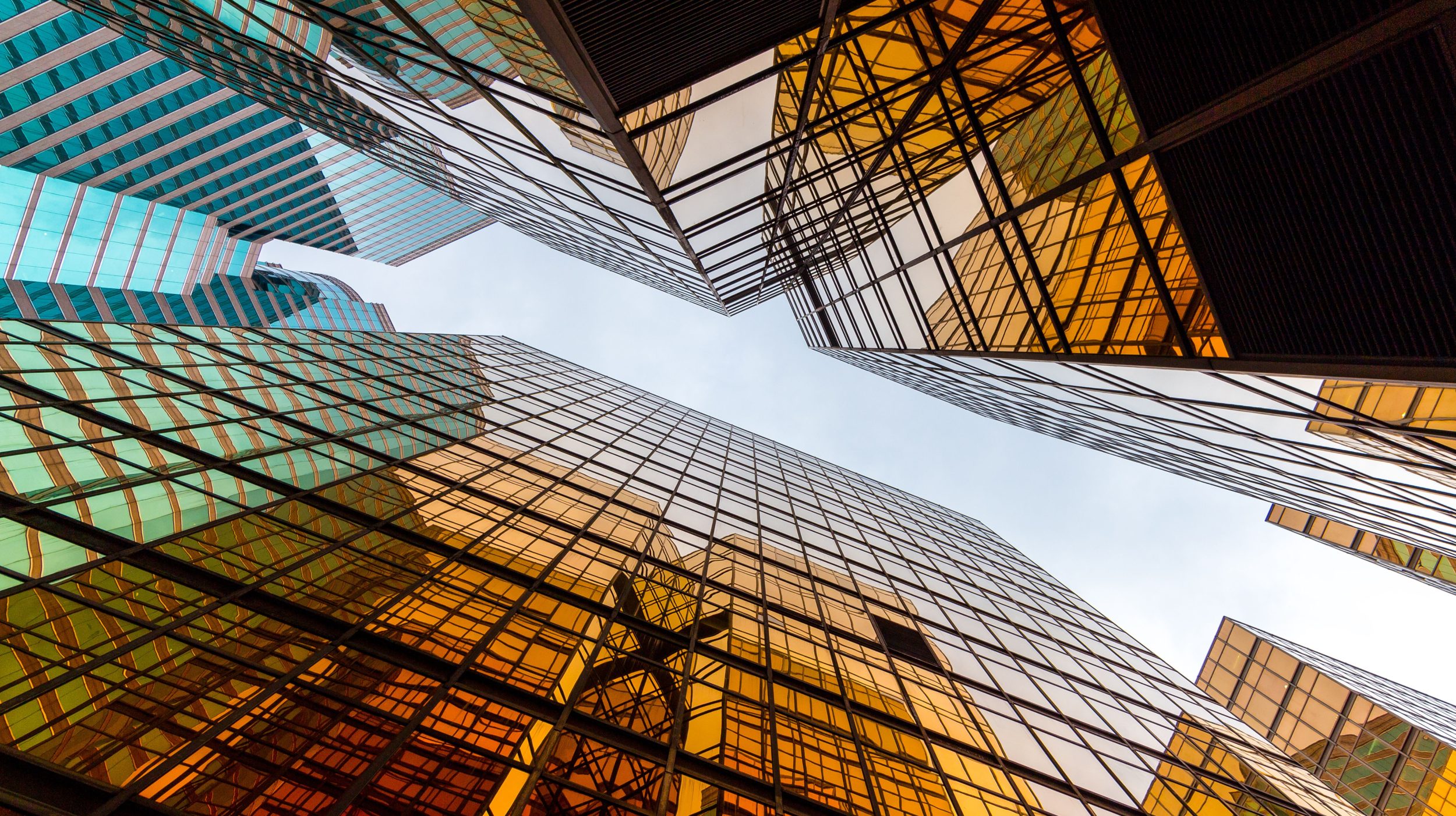 A low-angle view of modern skyscrapers with reflective glass windows in gold and turquoise hues, creating geometric patterns.